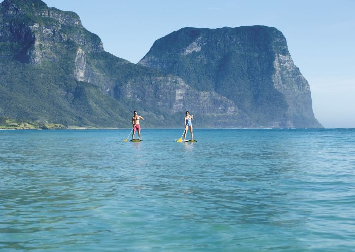 Couple stand-up paddleboarding at Lagoon Beach on Lord Howe Island with Mount Gower and Mount Lidgbird as the backdrop.