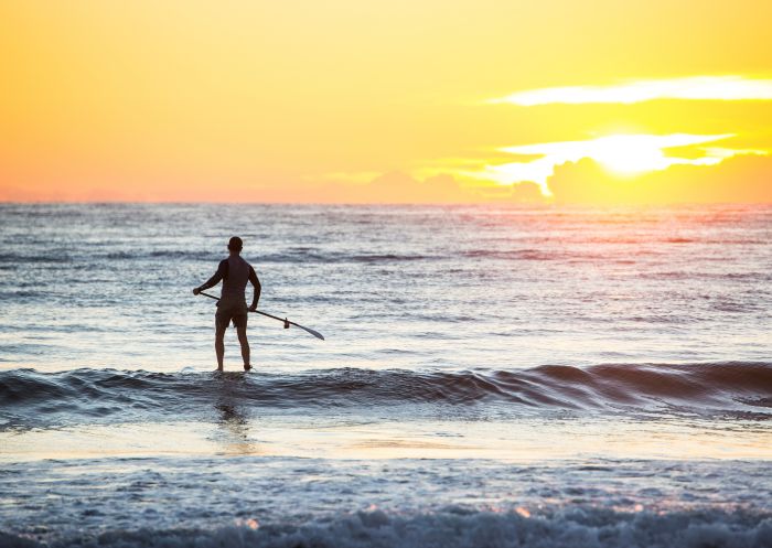 Man heading out for a morning of stand-up paddleboarding at Tathra Beach in Tathra, Sapphire Coast 