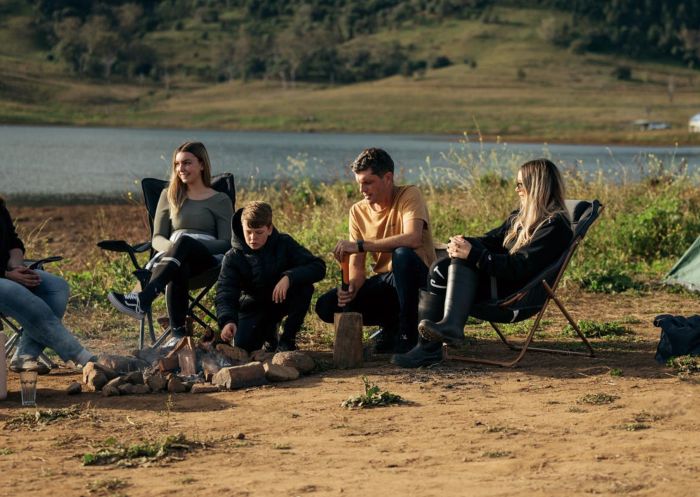 Family sits in camping chairs around a small campfire by the lake at Reflections Holiday Parks Lake Keepit, Tamworth