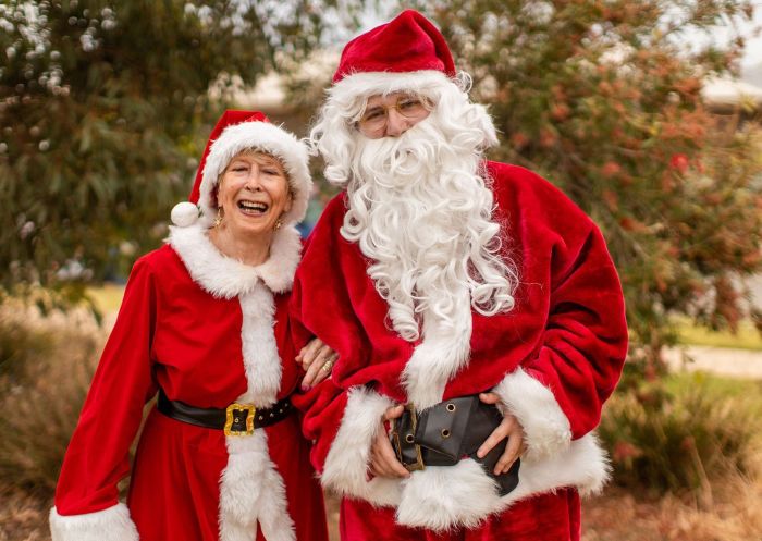 A woman and man in red santa suits in front of a tree at the Wagga Wagga Christmas Trail, Wagga Wagga