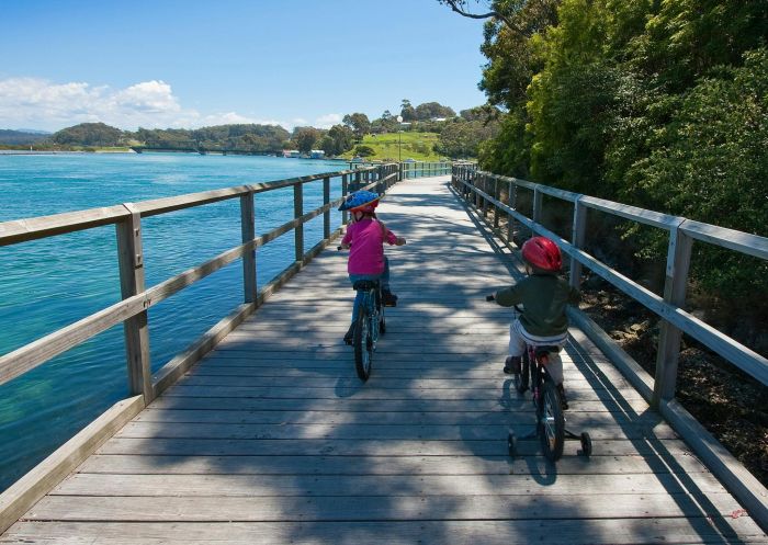 Children riding their bikes along a board walk by the water at Mill Bay Boardwalk and cycle-way, Narooma - Credit: Jon Poyner | Eurobodalla Tourism