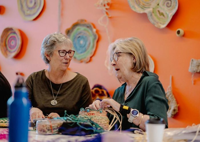 Two women having a conversation in front of an orange wall surrounded by woven baskets, Ngununggula Southern Highlands Regional Gallery, Bowral - Credit: Brian Rapsey