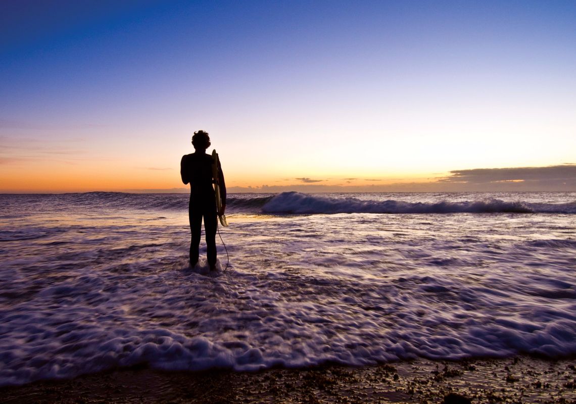 Ein Surfer beobachtet den Sonnenaufgang und das Brechen der Wellen am Angourie Point in der Nähe von Yamba.
