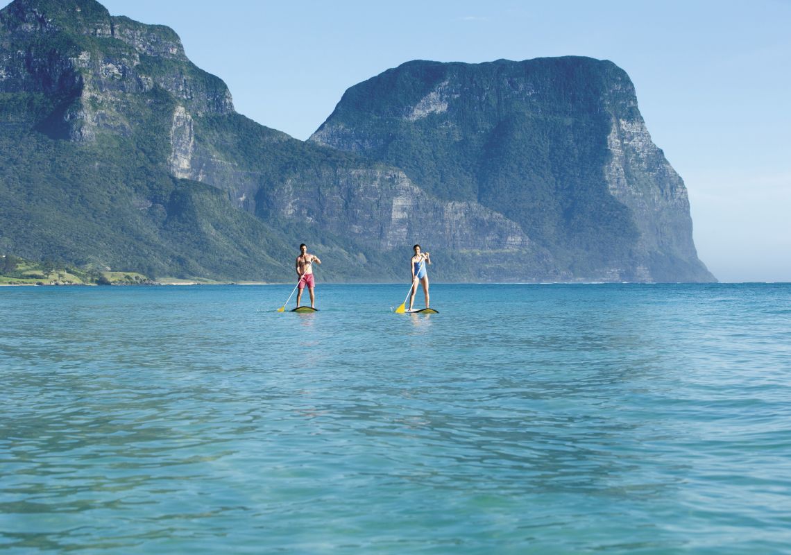 Couple stand-up paddleboarding at Lagoon Beach on Lord Howe Island with Mount Gower and Mount Lidgbird as the backdrop.