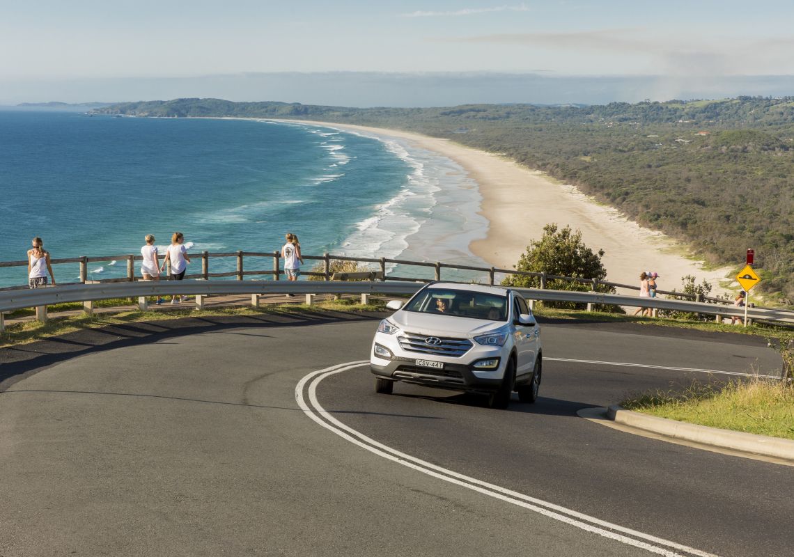 Car driving along the coast with Tallow Beach, Byron Bay in the background