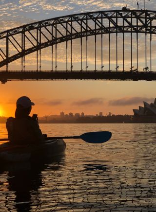 Silhouette of solo paddlers sitting on a calm Sydney Harbour with bridge and Sydney Opera House